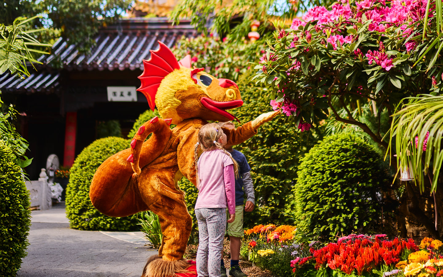 Zwei Kinder treffen Drache Pau Li auf einem Gartenweg zwischen blühenden Pflanzen und formgeschnittenen Sträuchern.