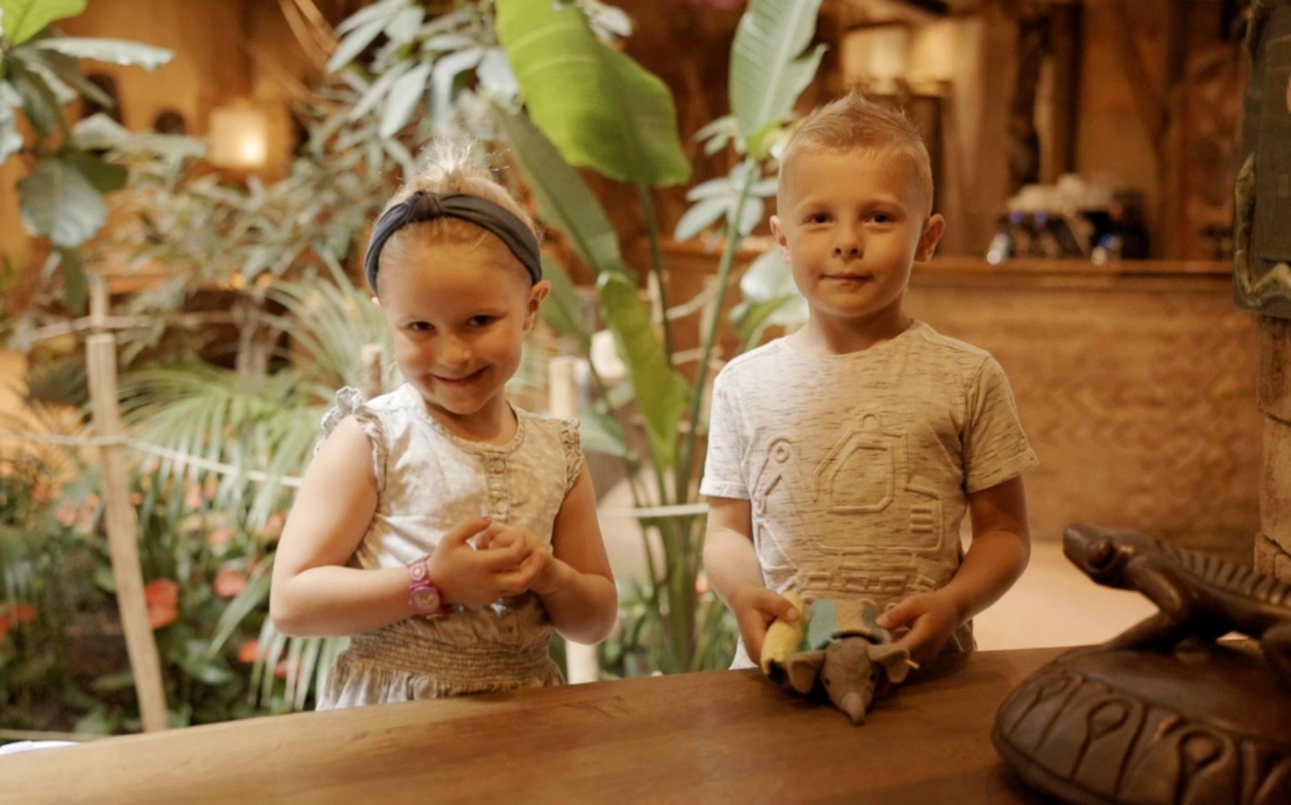 Zwei Kinder stehen an einem Holztresen vor tropischen Pflanzen und halten eine kleine Elefantenfigur in den Händen.